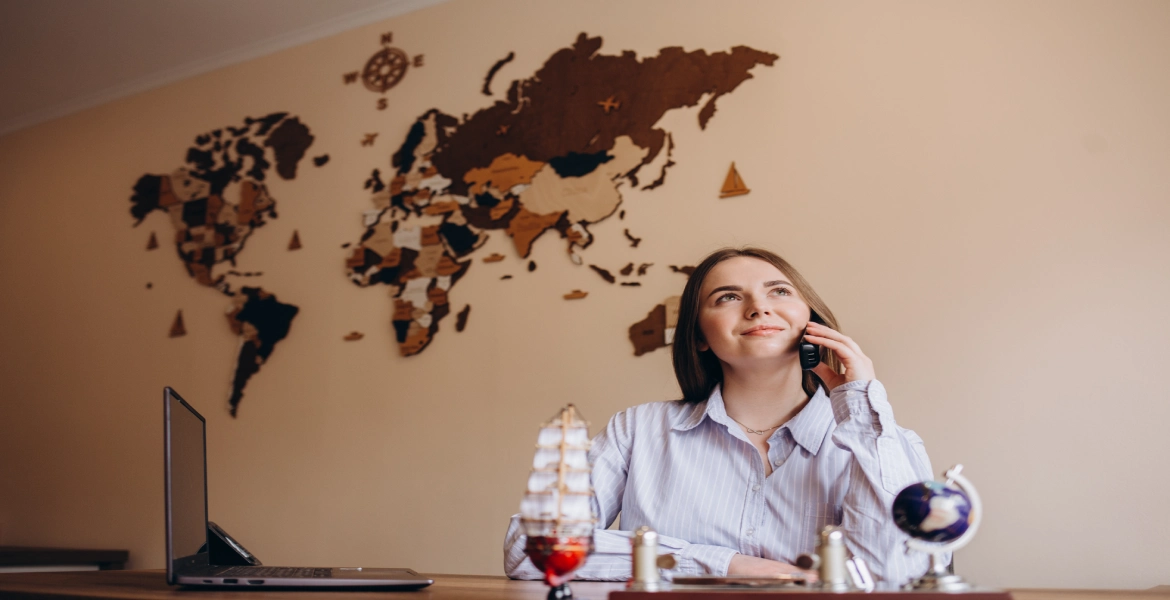 Mulher segura o telefone e está sentada em uma mesa com computador e um globo.